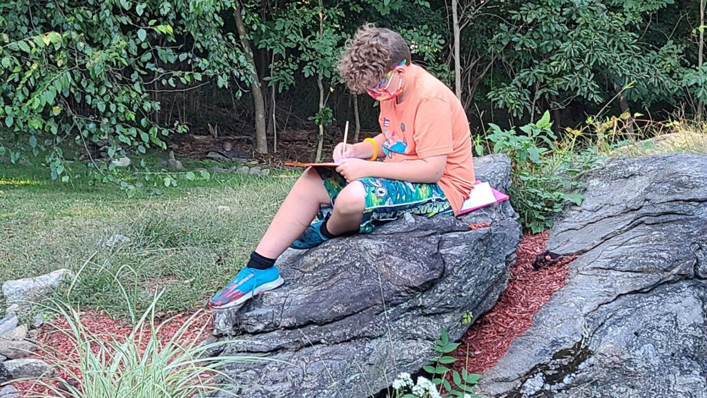 Child sitting on rock in wooded area, writing or drawing on a clipboard